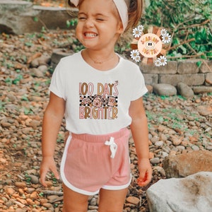 May include: A young girl wearing a white t-shirt with the text "100 Days Brighter" and a pink and white checkered pattern. She is also wearing pink shorts with white trim.