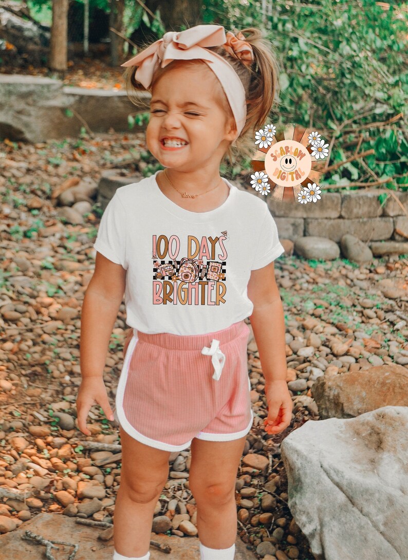 May include: A young girl wearing a white t-shirt with the text "100 Days Brighter" and a pink and white checkered pattern. She is also wearing pink shorts with white trim.