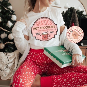 Op de afbeelding: Een wit T-shirt met een roze en witte afbeelding waarop staat "Santa's Hot Chocolate Made Fresh Daily Since 1962". De afbeelding toont een rode mok met marshmallows en een snoepstok. Het T-shirt wordt gedragen door iemand die een rode en witte stippenbroek draagt.