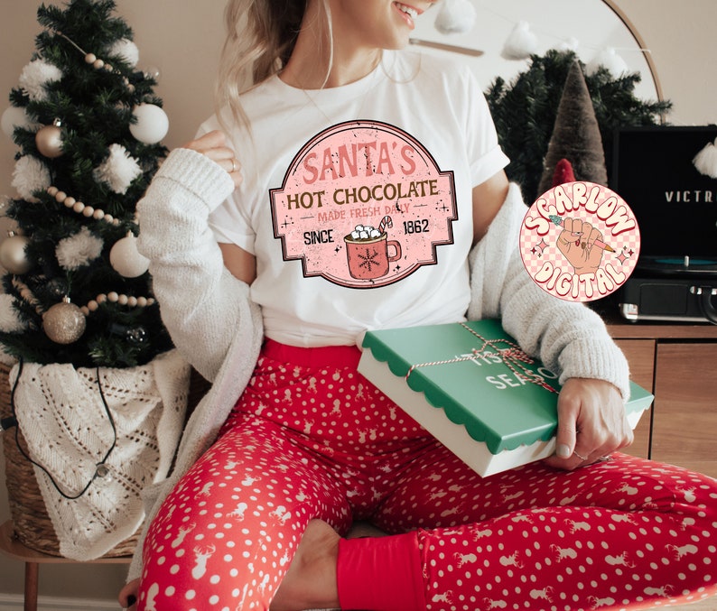 Op de afbeelding: Een wit T-shirt met een roze en witte afbeelding waarop staat "Santa's Hot Chocolate Made Fresh Daily Since 1962". De afbeelding toont een rode mok met marshmallows en een snoepstok. Het T-shirt wordt gedragen door iemand die een rode en witte stippenbroek draagt.