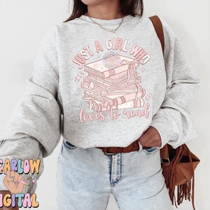 Puede incluir: Una sudadera gris con el texto "Just a girl who loves to read" en letras rosas. El texto est&aacute; rodeado de una pila de libros con flores rosas en el libro de arriba.