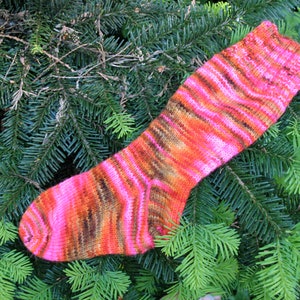 Puede incluir: Un calcetín de punto a rayas rosa, naranja y marrón. El calcetín está sobre un lecho de agujas de pino verdes.