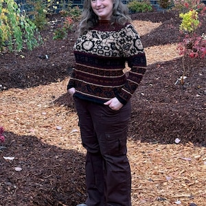 Può includere: Una donna indossa un maglione marrone e nero con un motivo a orologio e uccello. Indossa pantaloni marroni e si trova in un giardino con pacciame marrone.