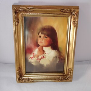 May include: A framed painting of a young girl with brown hair praying with her hands clasped together. The painting is set against a light yellow background with a soft glow around the girl. The frame is gold with ornate details.
