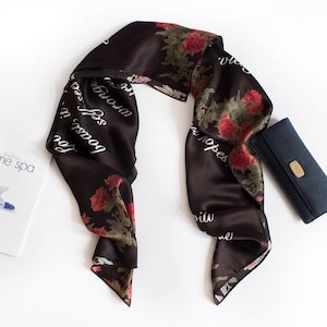 May include: A black silk scarf with a floral print and white text that reads "Good things in small doses for you". The scarf is folded in half and has a dark blue wallet next to it.