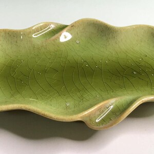 May include: An oblong ceramic dish with a wavy, organic edge and a light green glaze. The surface features a distinct crackle finish with darker green veining throughout, creating a textured appearance across the entire interior of the piece.