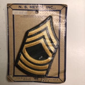 May include: A Master Sergeant military rank insignia patch with gold embroidery on a black background, mounted on a tan N.S. Meyer, Inc. display card. The card features the text N.S. MEYER, INC., 2PR MAST SGT, and INSIGNIA SPECIALISTS SINCE 1668.