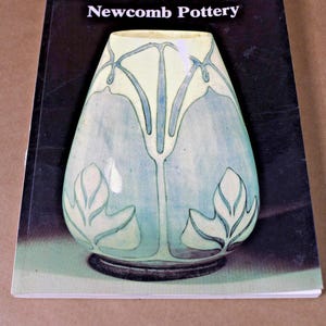 May include: A softcover book titled Newcomb Pottery featuring a cover image of a tapered ceramic vase with a light blue and cream glaze, decorated with stylized botanical line art and a dark base, set against a black background.