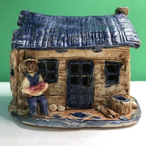 May include: A ceramic house figurine featuring a textured blue roof, a central door, and two windows. A figure sits on the left side holding a red object. The structure has a light tan finish with blue accents on the base, door, and roof trim.