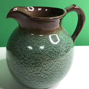 May include: A ceramic pitcher with a rounded body and a curved handle. The upper portion and handle feature a smooth, solid dark brown glaze, while the lower, spherical body displays a textured, speckled mint green and dark green finish.