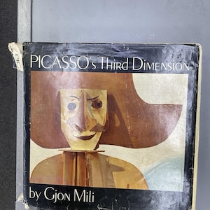 May include: A hardcover book titled Picasso's Third Dimension by Gjon Mili. The black cover features a central illustration of a stylized, tan-colored mask-like face with a wide-brimmed hat, set against a light background with black borders.