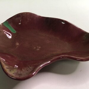 May include: A small, irregularly shaped ceramic dish with a glossy finish. The primary color is a deep, muted maroon or plum, featuring a distinct, bright green accent mark along one of the curved, raised edges and subtle yellow speckling.