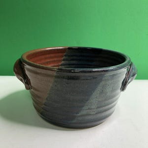 May include: A round ceramic bowl with two side handles, featuring a two-tone glaze design. One side of the bowl is a warm reddish-brown, while the other side is a deep, dark blue. The exterior surface has horizontal ridges throughout the body.