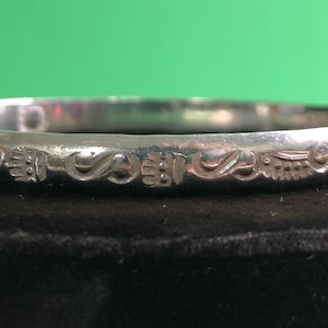 May include: A polished silver-toned bangle bracelet featuring a continuous band with engraved repeating patterns. The surface displays alternating motifs of small circular shapes, stylized leaf-like forms, and textured geometric elements.