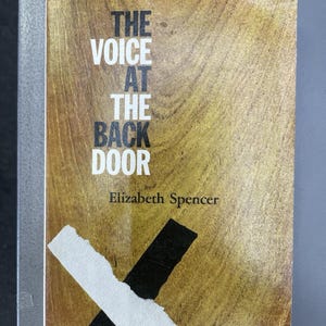 May include: A paperback book titled The Voice at the Back Door by Elizabeth Spencer. The cover features a wood grain pattern in shades of brown and tan, with black text and a large, overlapping white and black X shape across the bottom section.
