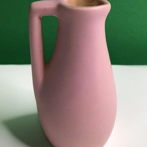 May include: A matte pink ceramic pitcher with a smooth, rounded body and a single integrated handle. The vessel features a simple, curved silhouette with a small pouring spout at the top rim, showcasing a consistent pastel pink finish throughout.