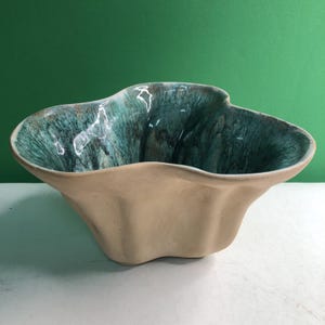 May include: A ceramic bowl with an irregular, wavy rim and a matte beige exterior. The interior features a glossy, deep teal glaze with dark, flowing streaks that create a marbled effect across the basin of the vessel, contrasting with the light base.