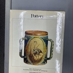 May include: A hardcover book titled Pottery from The Smithsonian Illustrated Library of Antiques, prepared by the Cooper-Hewitt Museum. The cover features a central illustration of a two-handled ceramic mug decorated with a flamingo in a marsh.