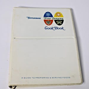 May include: A white Tupperware Four Seasons Cook Book with a ring binder mechanism. The cover features the text Tupperware, Four Seasons, Cook Book, and A GUIDE TO PREPARING & SERVING FOODS, along with four oval icons representing the seasons.