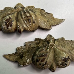May include: Two decorative metal hardware pieces featuring a relief design of two rounded fruits nestled among textured leaves. The items have a distressed, weathered finish with a muted beige and brown color palette showing signs of wear.