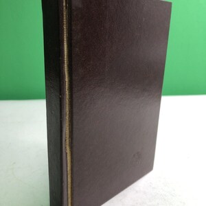 May include: A dark brown hardcover book standing upright. The book features a smooth, solid-colored cover with a visible spine edge showing light-colored binding material. The front cover has a subtle, embossed circular emblem near the bottom right.
