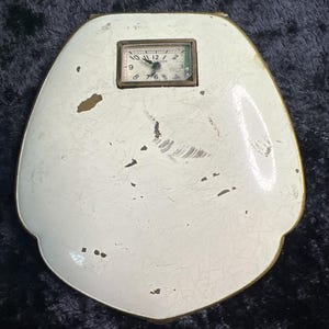 May include: A shield-shaped compact case with a white enameled finish featuring visible surface wear and cracks. A small rectangular analog clock with a white face and black numerals is embedded in the upper center of the white surface.