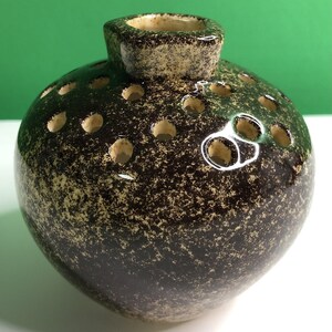 May include: A round ceramic flower frog vase featuring a dark brown and cream speckled glaze. The top surface is punctuated by numerous circular holes for arranging stems, and it has a small, raised central opening with a smooth rim.