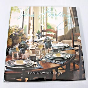 May include: A Vera Bradley cookbook titled Cooking with Friends featuring a photograph of a wooden dining table set for a meal with patterned plates, glassware, napkins, and floral arrangements in a bright, sunlit room with chairs.