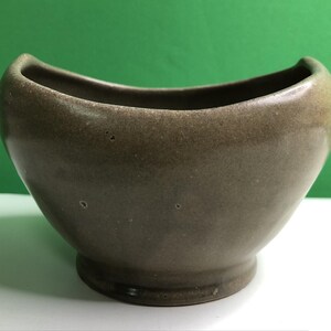 May include: A small, taupe-colored ceramic bowl with a smooth, glazed finish. The bowl features a distinct, curved, crescent-shaped rim that dips in the center and rises at both ends, supported by a circular pedestal base at the bottom.