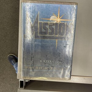 May include: A hardcover book titled Mission by Patrick Tilley with a blue, weathered-texture cover. The title is printed in large, bold letters with a gold star graphic above the letter i. The book is encased in a clear plastic protective cover.