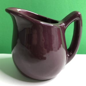 May include: A small, glossy, dark purple ceramic pitcher with a rounded body, a wide pouring spout, and a sturdy, curved handle. The surface of the pitcher has a smooth, reflective finish that catches the light, highlighting its deep, solid color.