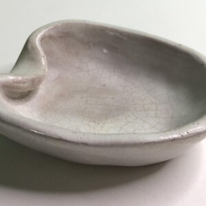 May include: A small, off-white ceramic dish with an irregular, organic shape and a raised, folded edge. The surface features a visible crackle glaze finish, creating a network of fine lines across the interior basin of the shallow, bowl-like object.