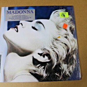 May include: A square vinyl record cover featuring a portrait of Madonna with platinum blonde hair and dark lipstick. The text reads MADONNA True Blue, LIVE TO TELL, and PAPA DON'T PREACH. A yellow sticker with the number 5 and an orange sticker are attached.