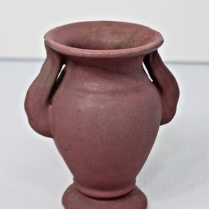 May include: A matte, dusty rose-colored ceramic vase with a wide, flared rim and two curved handles on the sides. The vessel features a rounded body that tapers down to a circular pedestal base, showcasing a uniform, textured, earthy finish.