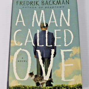 May include: A hardcover book titled A Man Called Ove by Fredrik Backman. The cover features a blue-toned sky background with a person in a dark blue jacket and cap standing in front of a green hedge, accompanied by a small cat in the foreground.