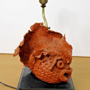 May include: A table lamp featuring a sculpted terracotta-colored fish base with textured scales and a large fin. The fish is mounted on a square black base with a brass-colored metal lamp socket and a curved neck extending from the top of the fish.