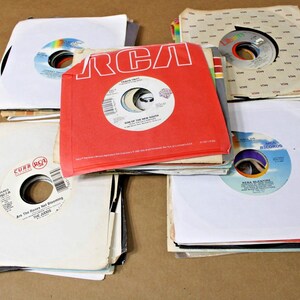 May include: A collection of five stacks of 7-inch vinyl records in paper sleeves. The center stack features a bright red RCA sleeve. Other sleeves are white with various record labels including Curb, MCA, and Liberty, showing artist names and song titles.