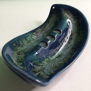 May include: A curved, crescent-shaped ceramic soap dish featuring a glossy glaze in deep navy blue and forest green. The surface displays a mottled, textured pattern with dark blue accents and three integrated drainage slots in the center.