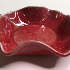 May include: A small, round ceramic dish with a vibrant red, glossy glaze and a textured, speckled finish. The bowl features a distinctively wavy, scalloped rim that creates a flower-like shape, with a smooth, flat circular base in the center.