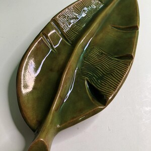 May include: A glossy, olive green ceramic dish shaped like a large leaf. The surface features a central vein running the length of the piece, with textured, horizontal ribbed patterns on alternating sections of the leaf blade and smooth areas.