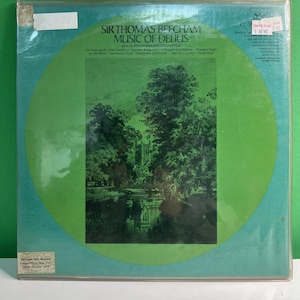 May include: A vinyl record album cover titled Sir Thomas Beecham Music of Delius by the Royal Philharmonic Orchestra. The cover features a green circular design with a central illustration of a church surrounded by trees and a pond.