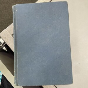 May include: A rectangular hardcover book with a solid, muted blue fabric cover. The book features a vertical spine on the left side with faint, worn gold-colored markings. The surface of the cover shows a subtle, uniform texture throughout.