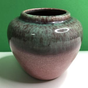 May include: A small, rounded ceramic vase featuring a glossy glaze with a color gradient. The upper portion displays a mottled mint green and dark brown finish, while the lower section transitions into a solid, textured dusty pink base.