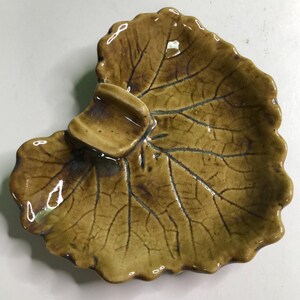 May include: A ceramic dish shaped like a large leaf with a glossy mustard-brown glaze and dark brown veining. The surface features a raised, curved indentation near the top edge, creating a distinct texture that mimics the natural form of foliage.