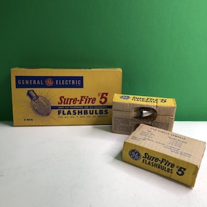 May include: Three yellow and brown cardboard boxes of General Electric Sure-Fire #5 flashbulbs. The largest box features blue text and a graphic of a flashbulb. Two smaller boxes show the General Electric logo and product information text.