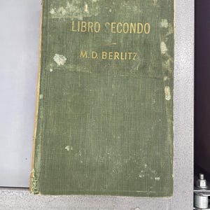 May include: A vintage hardcover book with a faded olive green cloth cover. The front cover features the text LIBRO SECONDO and M.D. BERLITZ printed in gold lettering. The edges of the cover show signs of wear and fraying, with visible light-colored patches.