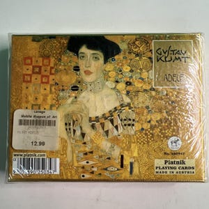 May include: A rectangular box of Piatnik playing cards featuring Gustav Klimt's Adele portrait. The gold-toned packaging displays the artwork, the text GUSTAV KLIMT ADELE, and a barcode label. The box is wrapped in clear plastic film.