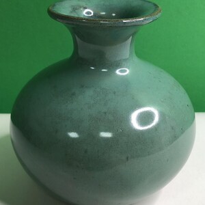 May include: A small, round ceramic vase with a glossy, teal-green glaze. The vase features a wide, bulbous body that tapers into a narrow neck, ending in a flared, circular rim. The surface shows subtle variations in color and texture.