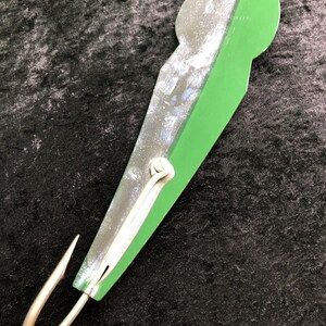 May include: A fishing lure featuring a long, tapered metal body with a split design of metallic silver and solid green. A single, curved, sharp metal hook is attached to the bottom end, and a small metal loop is located at the top for line attachment.