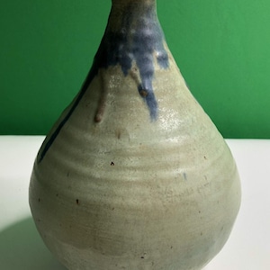 May include: A ceramic vase with a rounded body and a narrow neck. The surface features a textured, off-white glaze with horizontal ridges and a blue glaze drip pattern extending from the rim down the upper portion of the vessel's exterior.